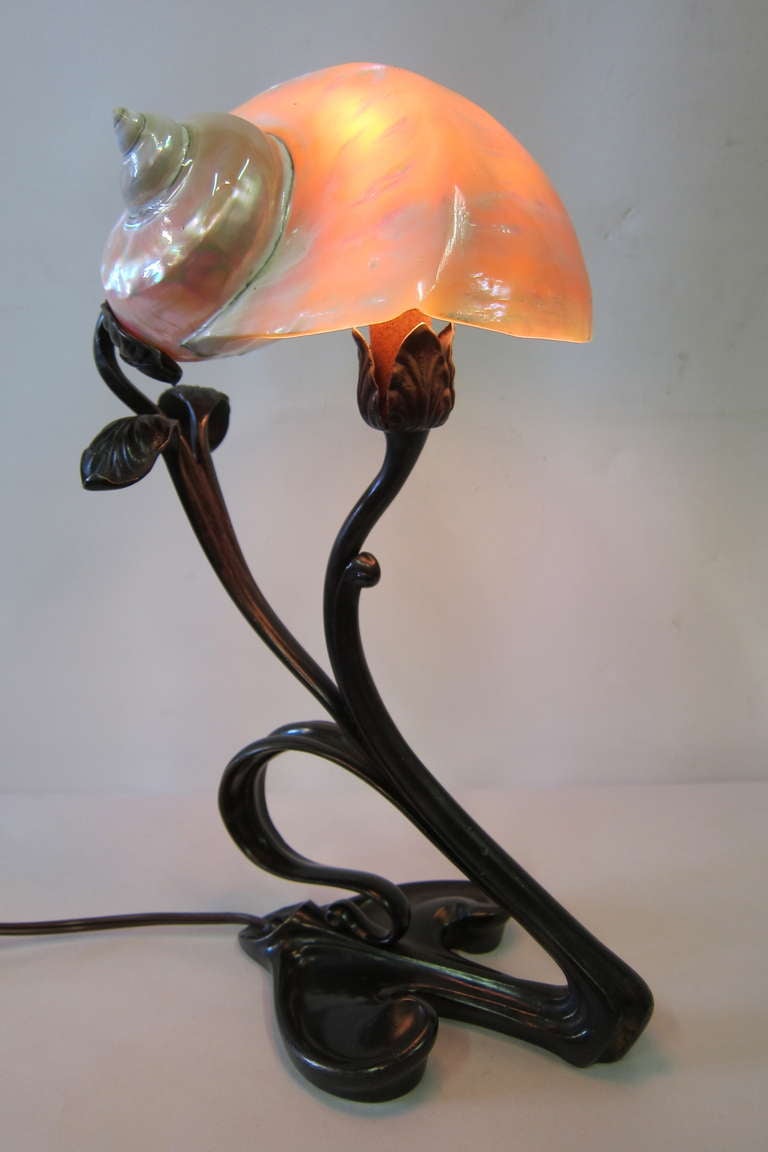 This bronze art nouveau lamp is designed with a stylized base that
celebrates the movement. The base has a rich dark chocolate patination that accents a lustrous conch shell shade. The shell possesses vibrant, iridescent colors that both highlight