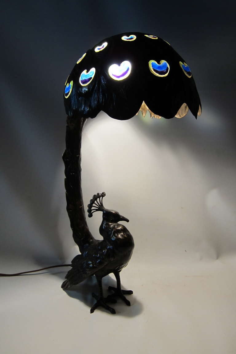 This bronze patinated art nouveau themed table lamp features a male peacock standing proud, displaying an upended canopy tail.
The tail is beautifully decorated with individually molded panels of art glass simulating splendid vibrant colors of
