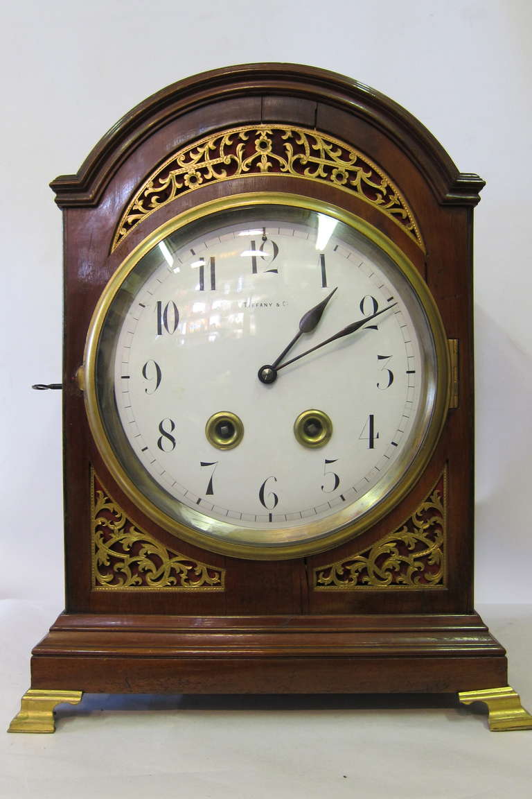 This impressive Tiffany mantle clock is designed in a rich cherry wood enclosure. This clock body is decorated with dore' gold bronze templets that adorn the front panel. These templets or stencils have a strolling design that is accented by a