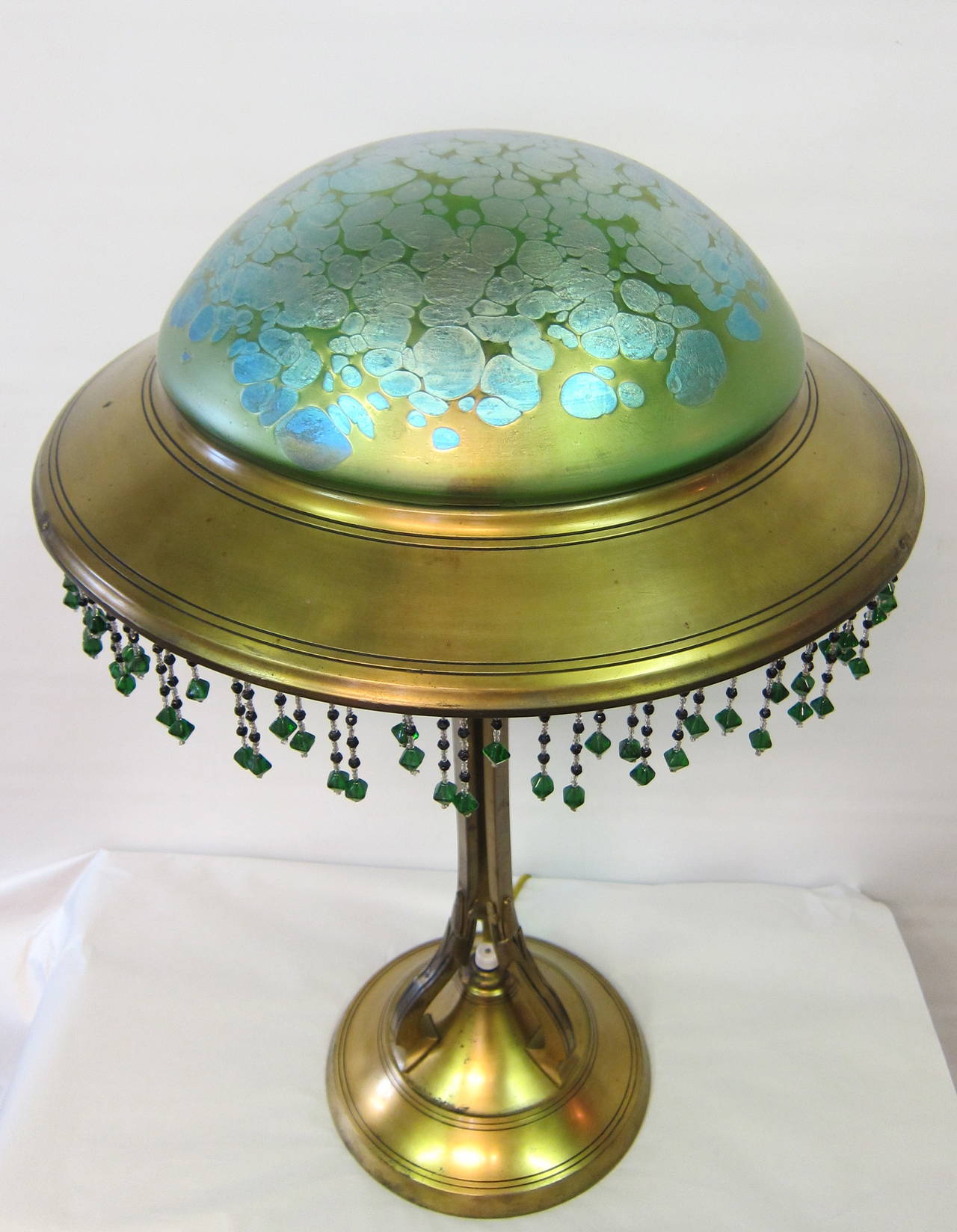 This early 20th century Austrian art nouveau table lamp is from the Loetz factory & has a blue oil stain pattern on green color dome shape shade.
The stunning 12