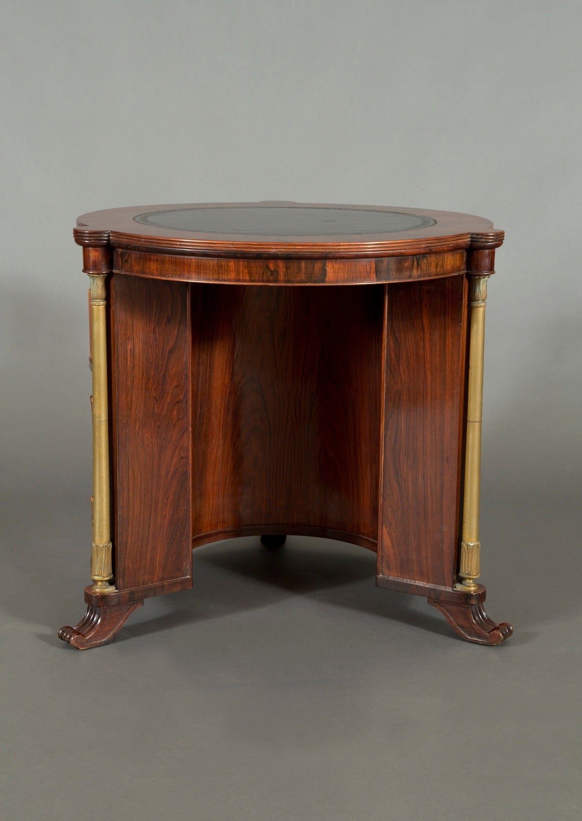 The crossbanded circular top centered by original blind tooled black leather. The kneehole base with a system of open bookshelf compartments divided by four gilt-brass columns with lotus leaf capitals. The whole raised upon four scrolled and molded