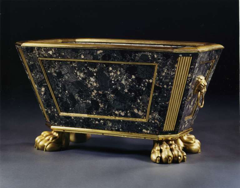 Of sarcophagus form, the paneled sides with gilt bronze molding, surmounted by a replaced cushion mold, the canted angles with reeded bronze panels, the two short sides mounted with lion-mask ring-handles, the whole raised on four gilt bronze paw