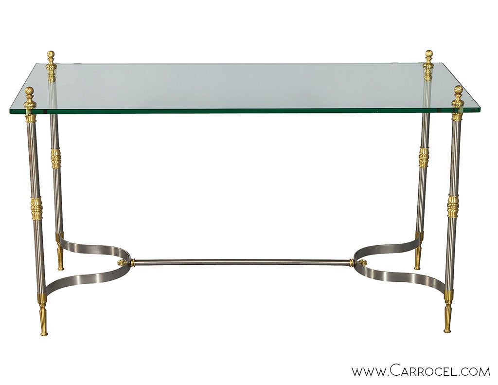 Console table reflecting the design style of Maison Jansen. The frame is made of brushed steel accented by brass details. Round finials secure the thick glass top to the frame.