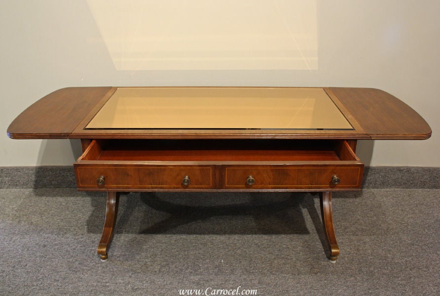 This is a solid mahogany antique coffee table with a satinwood banding and solid brass hardware. It features an original glass top and practical fold-down drop leaves on each end. Adding to its practicality, is a spacious drawer in the middle for