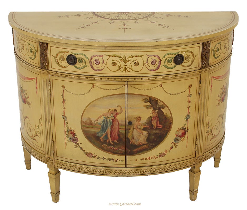 Here we have a masterfully hand-painted demi-lune commode made in America.  It has been finished in a warm cream colour and finished off with a beautiful painted classical scene in the middle cabinet door.  The detailing on this piece is magnificent