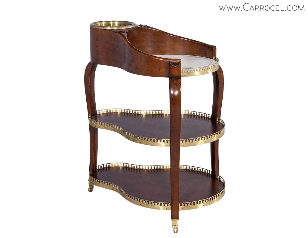 A three-tiered bar cart with the richness of a mahogany frame, brass accents and a white marble top. Three tapered, slightly sabered legs with brass casters support three serpentine shelves, the top flowing seamlessly into an inbuilt bottle holder