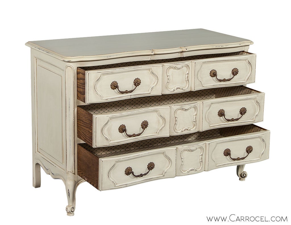 A beautiful Louis XV French Provincial style commode in a distressed ivory finish with straight sides and a graceful serpentine front consisting of three drawers with recessed panel fronts, molded outlines and two brass rosette loop handles each.