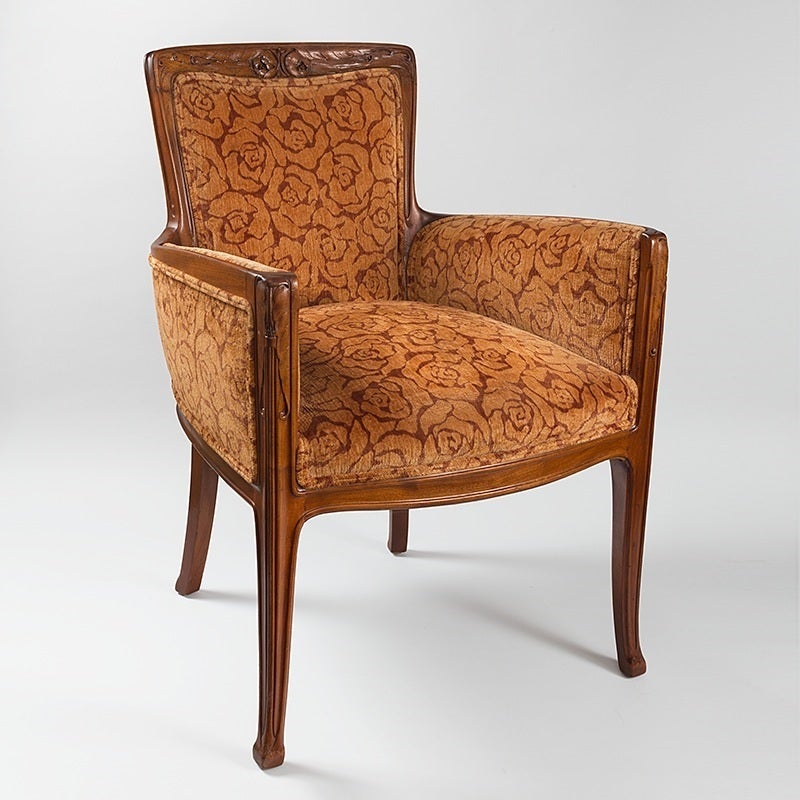 A French Art Nouveau bergère by Louis Majorelle. An upholstered armchair with an exposed mahogany wood frame. The curved fluted crest is decorated with carved stylized rose blossoms and leaves above a padded tapered rectangular back over a