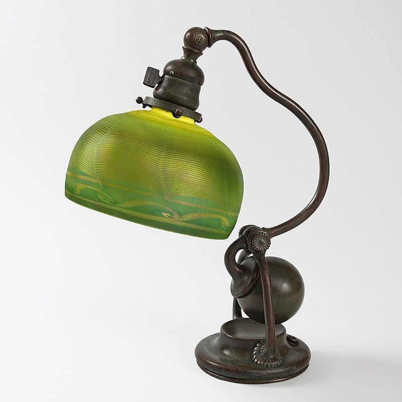 A Tiffany Studios New York patinated bronze and Favrile glass desk lamp featuring a decorated green “Damascene” shade with iridescent decoration atop a single arm counter balance base, circa 1900.

A similar shade and identical base are pictured