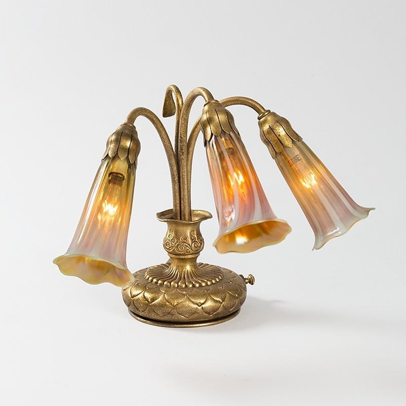 A Tiffany Studios New York glass and bronze three-light “Lily” piano lamp, featuring three iridescent gold Favrile “Lily” shades suspended from a gilt bronze decorated base. Circa 1900.

A similar lamp is pictured in: Tiffany Lamps and Metalware: