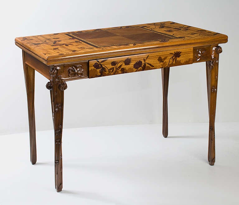 A French Art Nouveau games table by Louis Majorelle, featuring an inlaid marquetry top and carved legs and skirt. The marquetry decoration features stems, leave and, flowers around a central, bordered section. There is also  marquetry decoration on