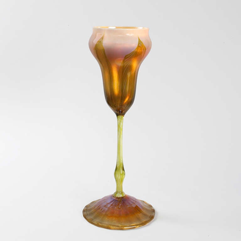 This flower form Favrile glass vase, by Tiffany Studios New York, features an organically-shaped, cream-colored upper portion, beautifully decorated with pulled feathering decoration in iridescent green and golden hues that are so bright, so