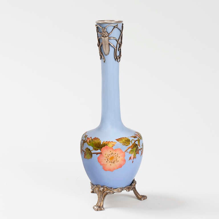 A French Art Nouveau silver and enamel vase by Eugène Feuillatre. The vase is decorated with leafed branches holding pink and gold cloisonné flowers and buds against a sky blue background. It rests on a braided silver stand with three carved feet.