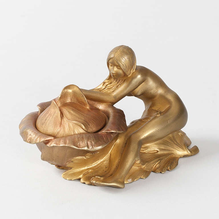 A French Art Nouveau gilt bronze salt cellar by Maurice Bouval formed from a female nude perched delicately next to a blossom, designed to house the salt.  With her hands the woman is gently pulling back the petals of the flower, inviting the viewer