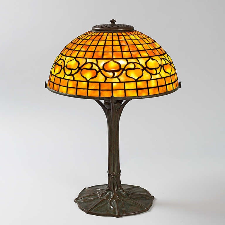 A Tiffany Studios New York “Acorn” glass and bronze table lamp. The shade features an orange and yellow acorn vine against a mottled orange glass geometric ground and sits atop a bronze three-armed “Penguin” base. Circa 1900.

A similar shade and