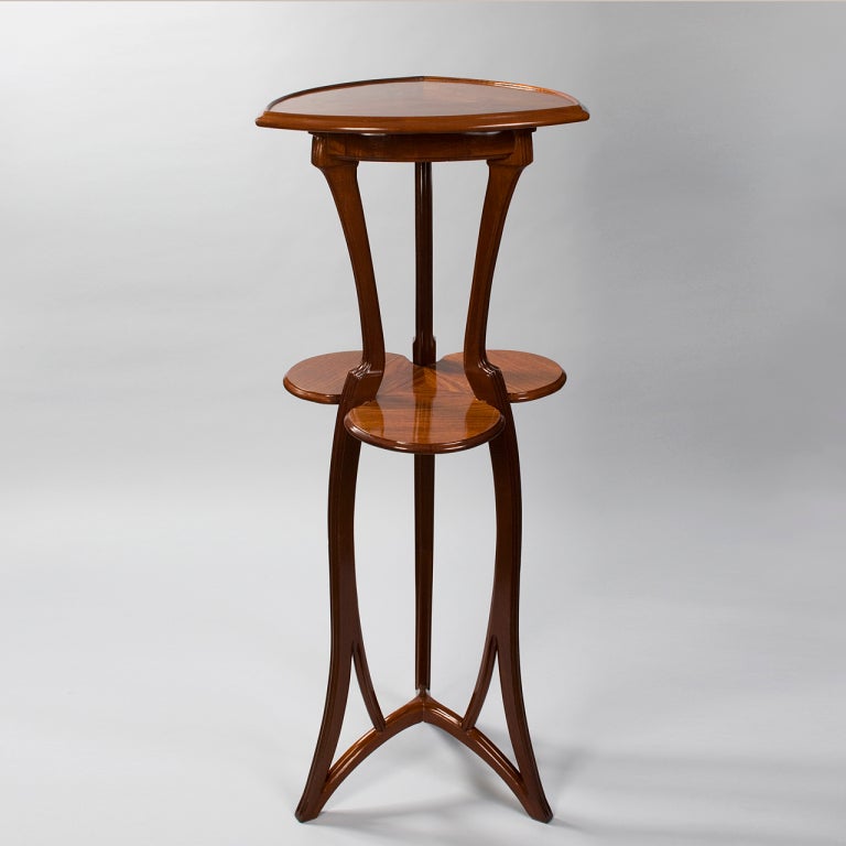 A French Art Nouveau two-tier wooden pedestal by Louis Majorelle, featuring a triangular top, a clover-shaped second tier and three sinuous, carved supports. 

A similar table is pictured in Majorelle - Nancy: décorations d'INTÉRIEURS: meubles,