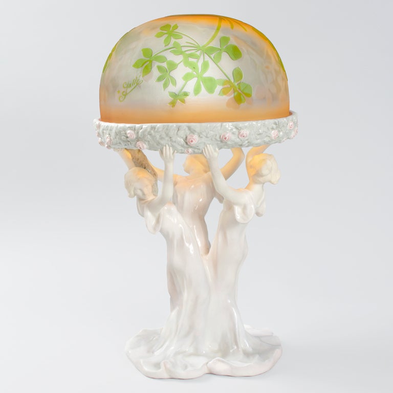 An Art Nouveau cameo glass and Vienna Faience glazed figural lamp by Emile Gallé and Podany. The porcelain base is formed from three dancing women with outstretched arms holding a garland of pink roses. The blossoms echo the elegant dome-shaped