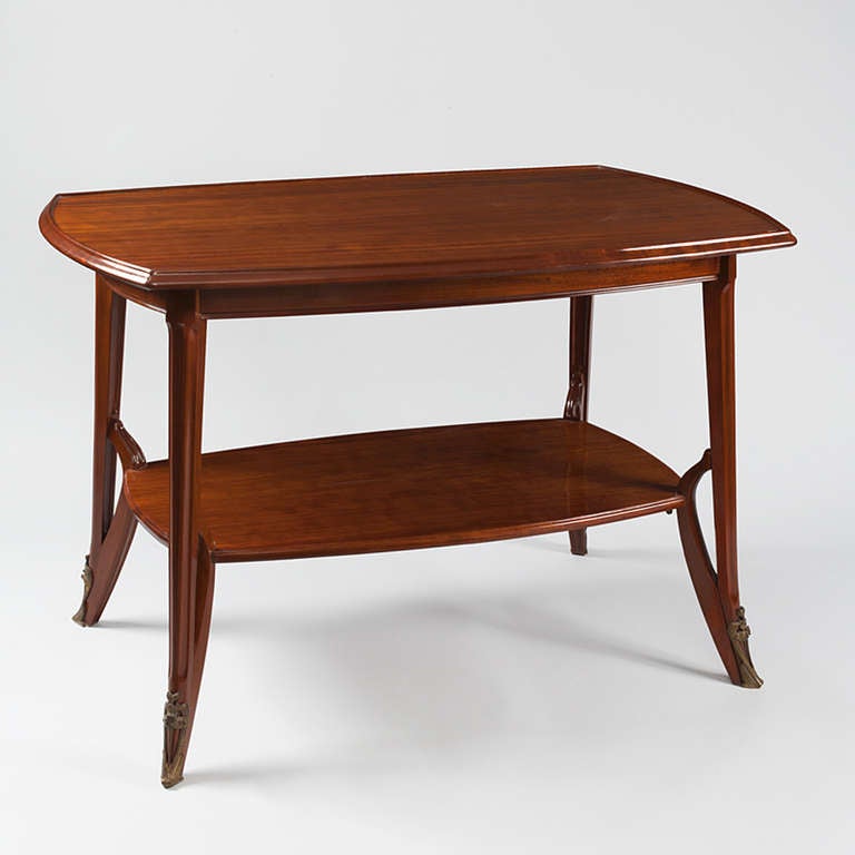 A French Art Nouveau mahogany center table by Louis Majorelle, the rounded top above a slightly bowed frieze, over downswept tapering channeled legs joined by a conforming undertier, ending in attenuated foliate cast sabots.  Circa 1900.

A