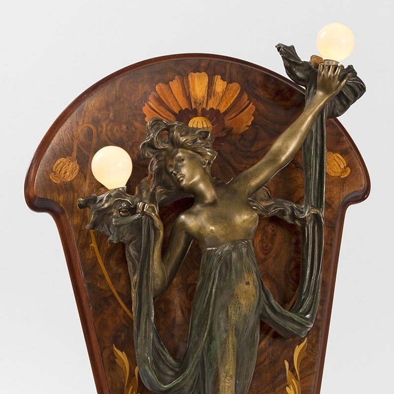 A French Art Nouveau bronze draped female nude in the form of a two-light sconce by Georges Flamand. The draped maiden, in a two-color patina, is supporting two lighting elements mounted on a wooden marquetry easel in a floral and thistle motif,