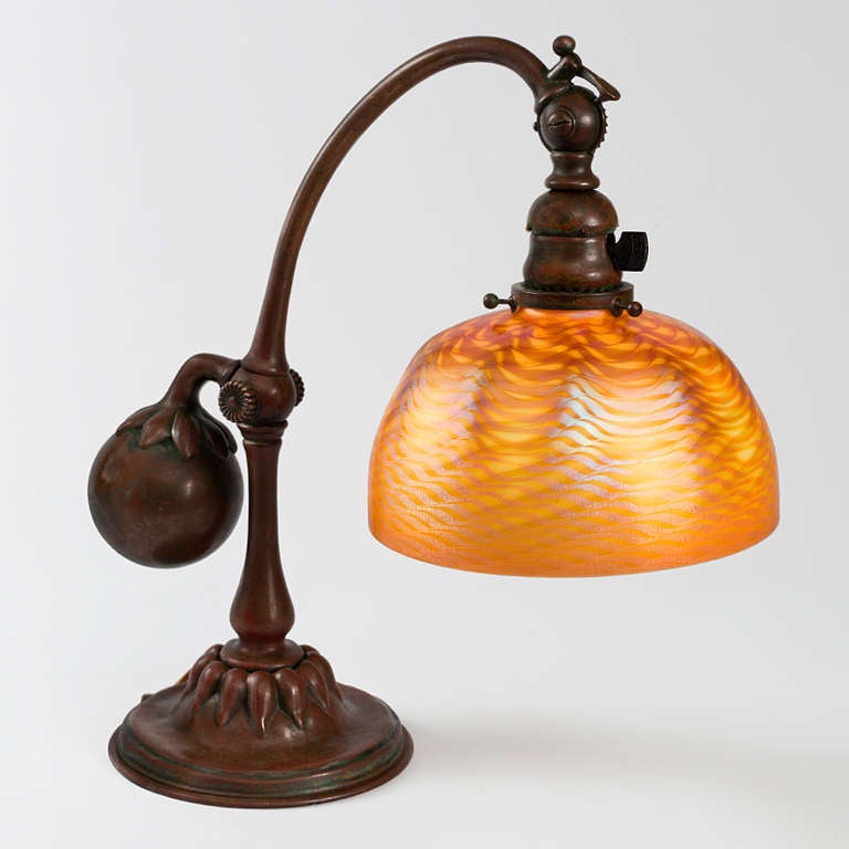 A Tiffany Studios New York Favrile glass and patinated bronze desk lamp with an iridescent gold Favrile glass “Damascene” shade suspended from a patinated bronze “Counterbalance” base, circa 1900.

A similar shade and base are pictured separately