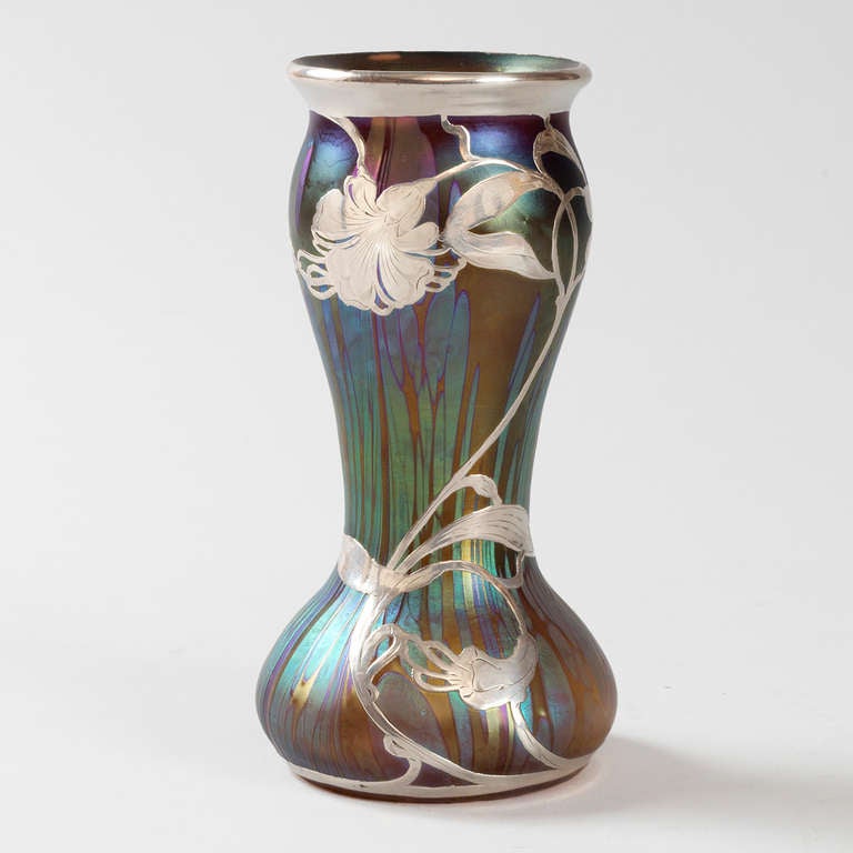 An Austrian Jugendstil iridescent glass vase with turquoise blue streaks over  a dark green ground by Loetz. The vase is further decorated with sterling  silver overlay in a floral motif. Circa 1900.
(MG Inventory #G15797)