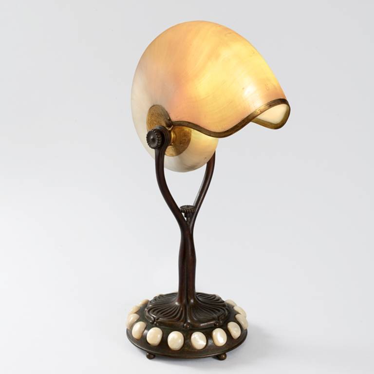 A Tiffany Studios New York “Nautilus” table lamp. This lamp features a nautilus shell shade set within a gilt patinated bronze tendril vine base. The base is further decorated with iridescent glass cabochon jewels. A similar lamp is pictured in:
