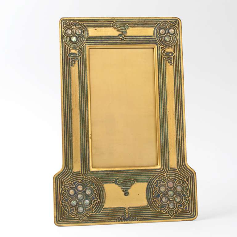 A Tiffany Studios New York  “Abalone” frame.  The frame is gilt bronze and decorated with an etched pattern.  The corners are further ornamented with thin abalone shell disks. (T-14879)

A similar pictured frame is pictured in: “Tiffany Lamps and