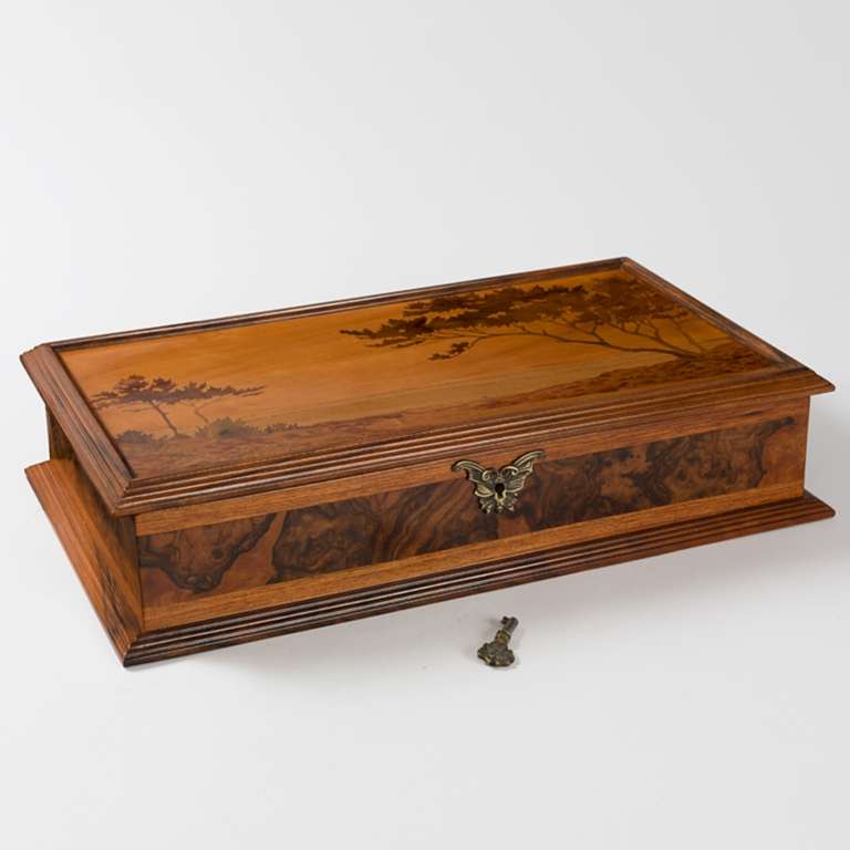 A French Art Nouveau lidded box with marquetry decoration in a landscape motif by Emile Gallé. The detailed marquetry work makes this piece appear more painted than carved. Galle’s landscape entices the eye to follow its sweeping line and invites