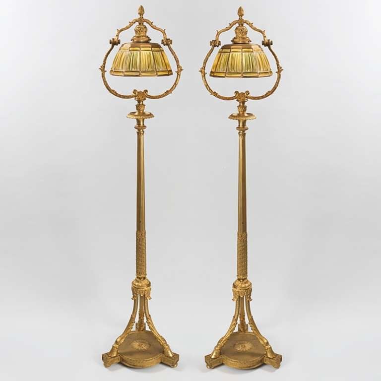 A pair of Tiffany Studios New York gilt bronze and “Linenfold” floor lamps. Each gilt bronze standard is raised on hoof feet and highly ornamented in a vegetal motif. Each shade formed from amber Favrile glass “Linenfold” panels is suspended within