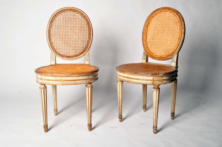 These round back chairs are from France and made from oakwood and rattan, circa 1900. A popular design during the 19th century, these round back chairs with original ivory paint feature airy caning on the back and seat. The legs are fluted and