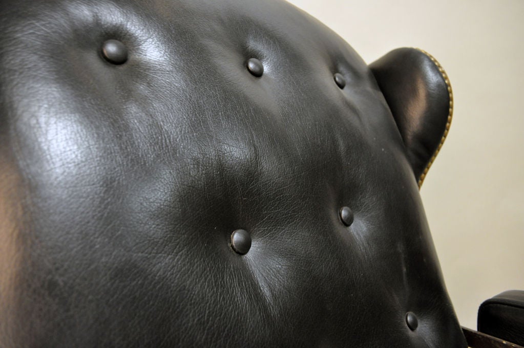 The seat, arms, and tufted back are upholstered in black leather, as well as the wings--which also feature brass upholstery tacks. The scrolling foliate-form finial rests atop the metal frame, which terminates in wheeled feet.