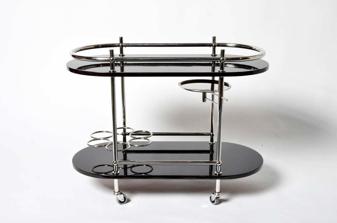 This contemporary two-tier bar cart features oval shaped high-gloss veneer trays on a tubular chrome frame. The shiny metal continues throughout to form a railing on the upper shelf, as well as a circular six-bottle rack below. One of the legs is