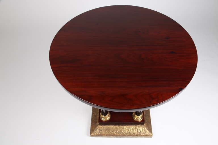 This handsome high-gloss table features a circular top supported by a central square pillar. Neoclassical influences can be seen in the added brass mounts and architectural elements: Four ebony colored columns that terminate in brass torus-form