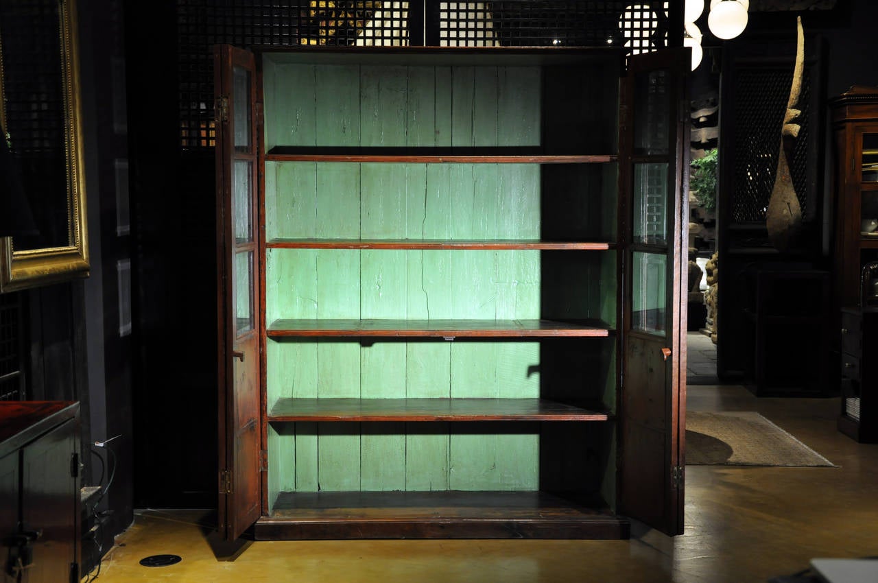 This charming teak bookcase has a painted red-brown exterior and was made in Rangoon, Burma in the Anglo-Indian style. Behind the double-hinged glass doors lies a soft aqua-green interior, typical of the time. This is an unusually animated and