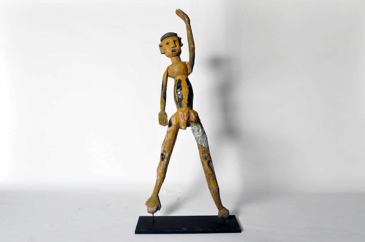 This large figure was carved by the Sukuma people. It is painted in yellow and black, and has rudimentary sculpted features. Both arms rotate, and a sheet of metal has been wrapped around the upper left thigh.