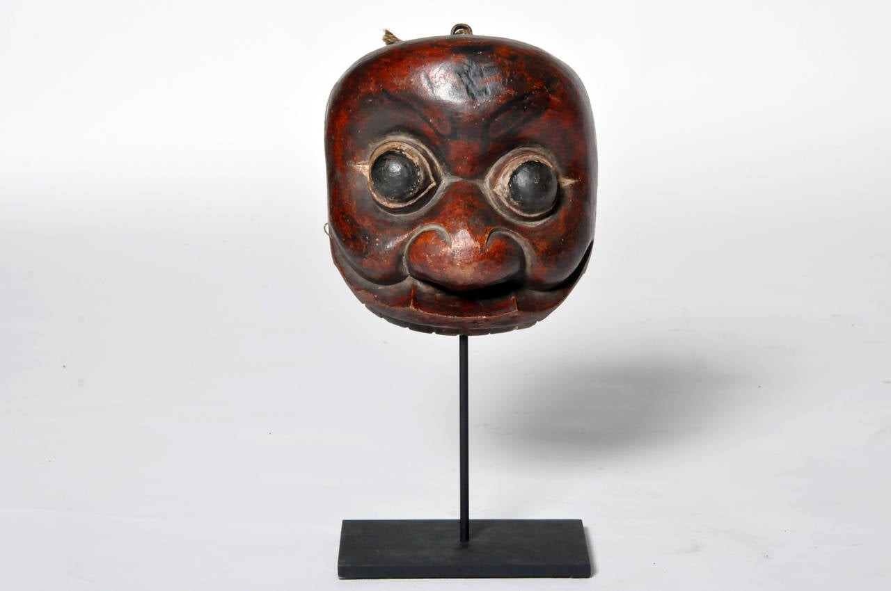 Muted slightly by a well-aged patina, the deep red lacquer and rounded features give this mask a dark and mysterious air. It is raised on a contemporary metal stand.