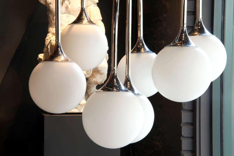 Descening from a circular canopy, the seven chrome arms terminate in flared bells that connect to glass spheres. When illuminated it provides a soft, off-white glow.