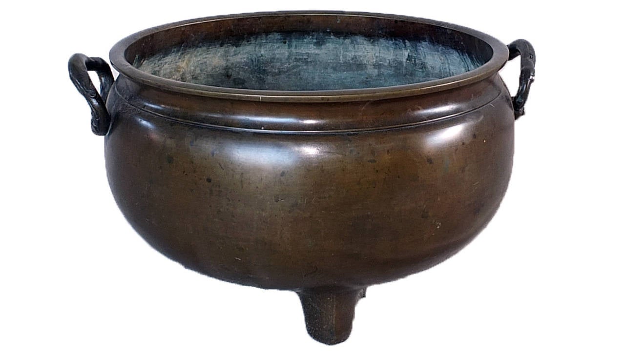 The wide-mouth bowl is flanked by decorated handles and raised on three cylindrical squat feet. The smooth surface has developed a handsome, lightly speckled patina.