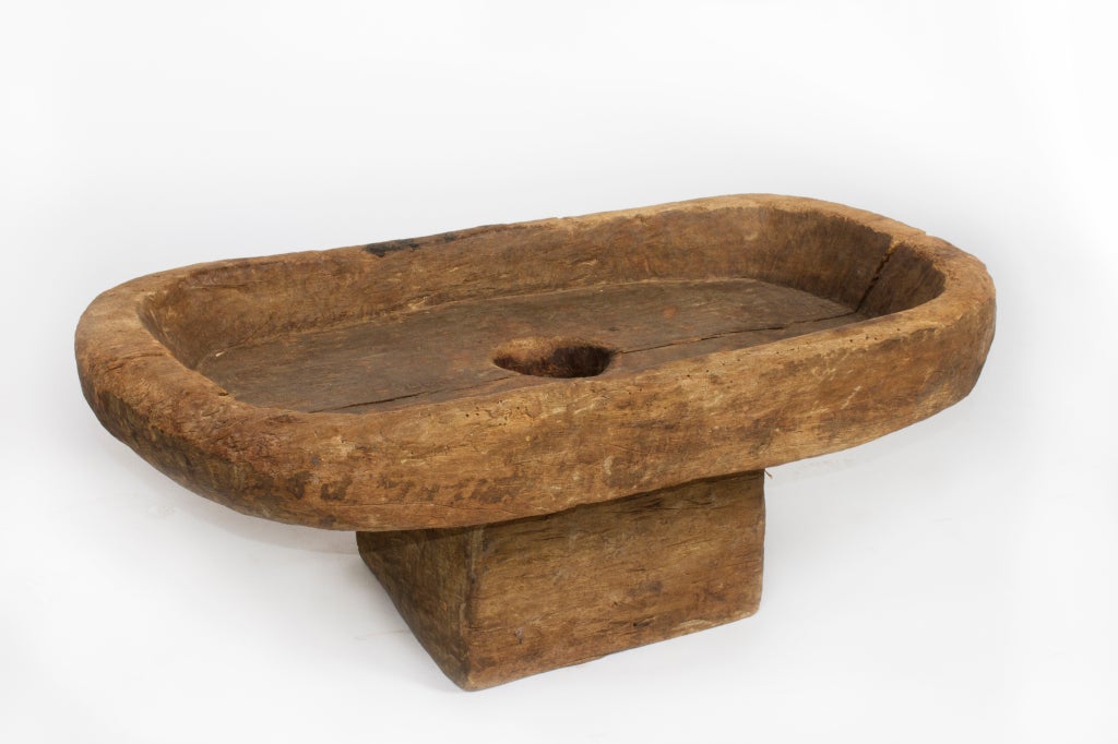 Mortar-and-pestle rice pounders like this are used to de-hull grains. This low, oval-shaped mortar is raised on a rectangular pedestal base. The wide top has raised edges to create a basin-like platform around the single hole at its center. Rice