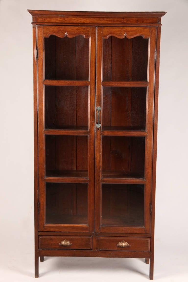 This stately British Colonial display cabinet is from Burma and is made from Teak wood. It dates to the early 20th Century. It features tall display windows and 2 bottom drawers for additional storage. It is ideal for art books. Teak Wood was