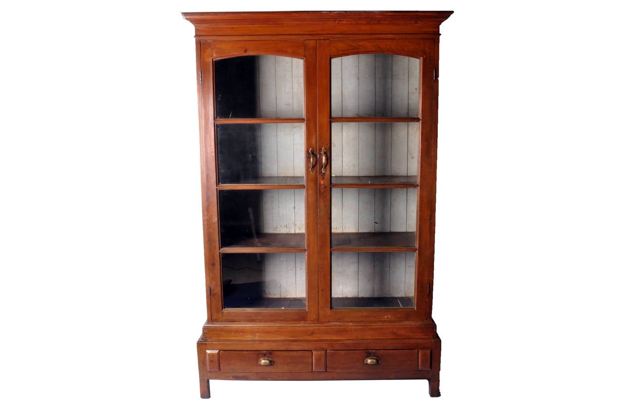 This gorgeous British Colonial display cabinet was made in Rangoon, Burma from teakwood and dates to the early 1900s. This display cabinet features shelves, two drawers and beautifully aged patina.