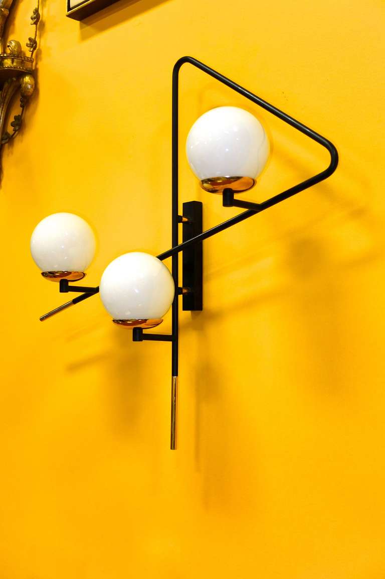 French 1950's asymmetrical triangular form black matte painted brass tube wall light with three white glass ball globe shades which rest on round brass disk bobeches.