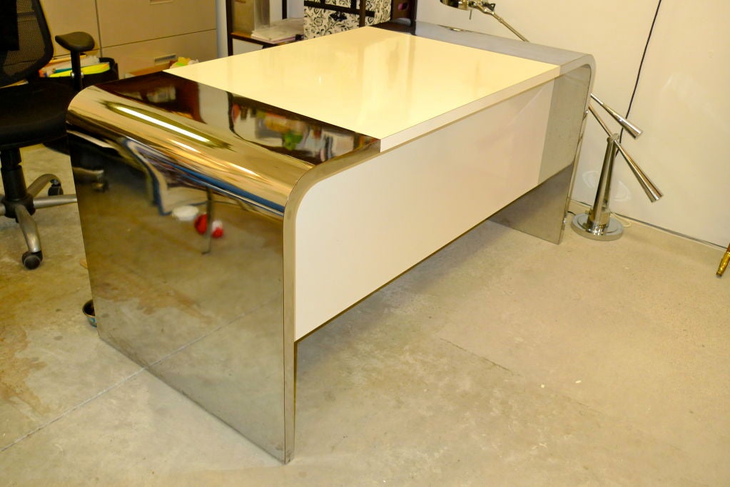 A full size desk in waterfall form completely clad in satin white and brilliant chrome laminate.  Single top center drawer and a low single shelf.  Knee-hole space sufficiently ample to allow for a rolling two drawer filing cabinet. Guest facing