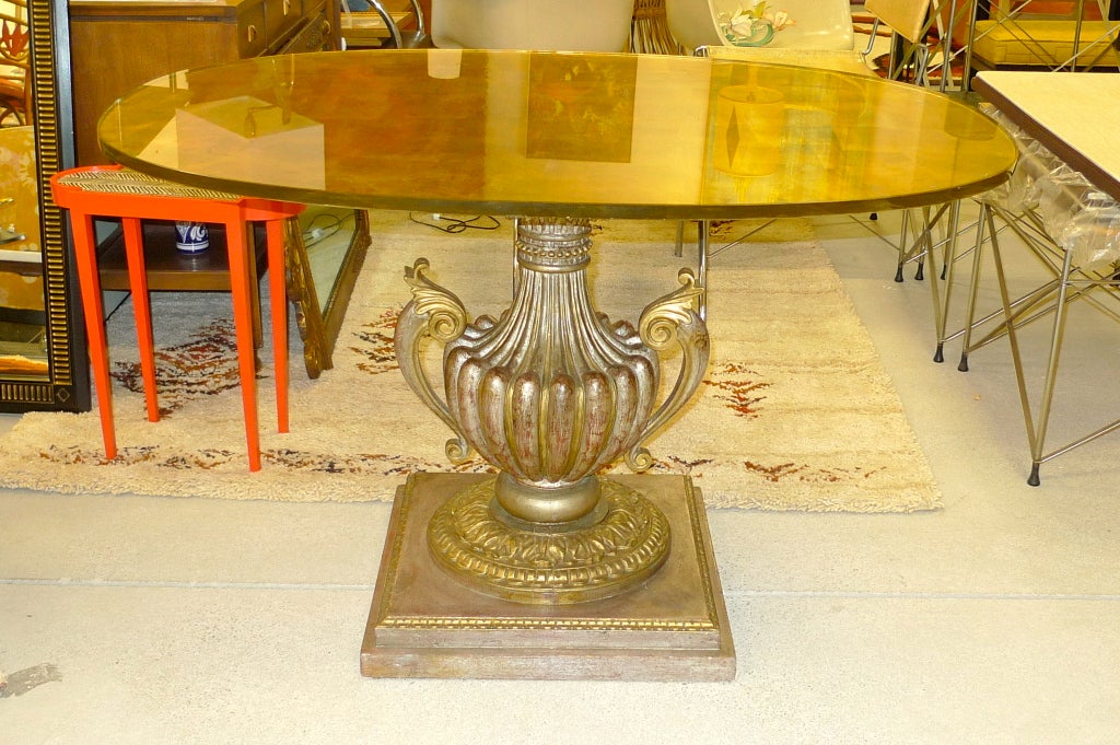 Gracious center table with an elaborately carved two-handled fluted urn-form pedestal base terminating in a round foot mounted on a classical square plinth.   The oval shaped glass top (1/2 inch thickness) is gilded on the reverse.