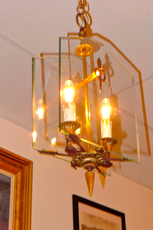 This mid-century Italian lantern pendant has elements of Fontana Arte style, notably geometric glass panels mounted in a bronze structure.  However, this fixture has elements of the Directoire style, namely flame tor hes, laurel rings and crossed