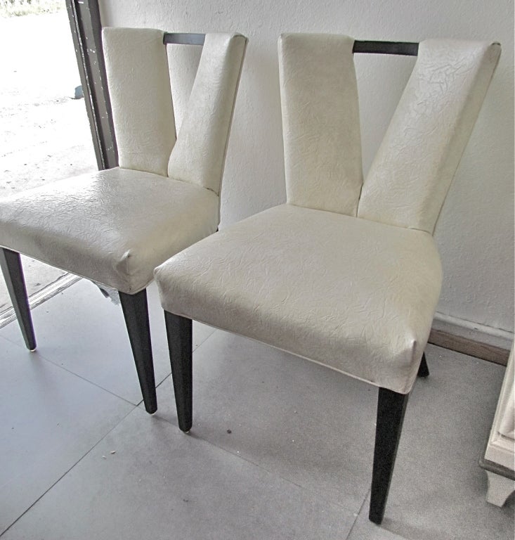 Va-va-Voom!  Four elegant Paul Frankl 'Corset' chairs with V-back forms and a wood cross-bar detail.  Black lacquered  frames and textured white vinyl upholstery.
Height 32.5”. Width is ~20” wing tip to wing tip. Depth ~20”. Seat height