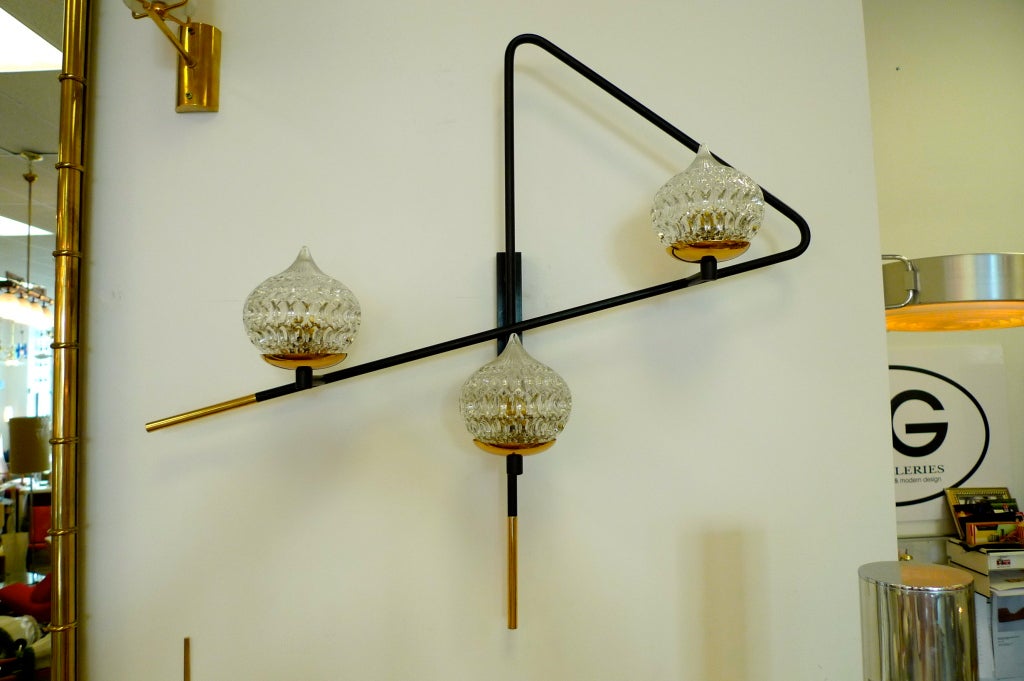 French 1950's triangular paperclip form black matte painted tube rod wall light with three onion form molded glass shades which rest on round brass disk bobeches, and long brass finials.

Able to be hardwired or mountedm with a power cord. We have