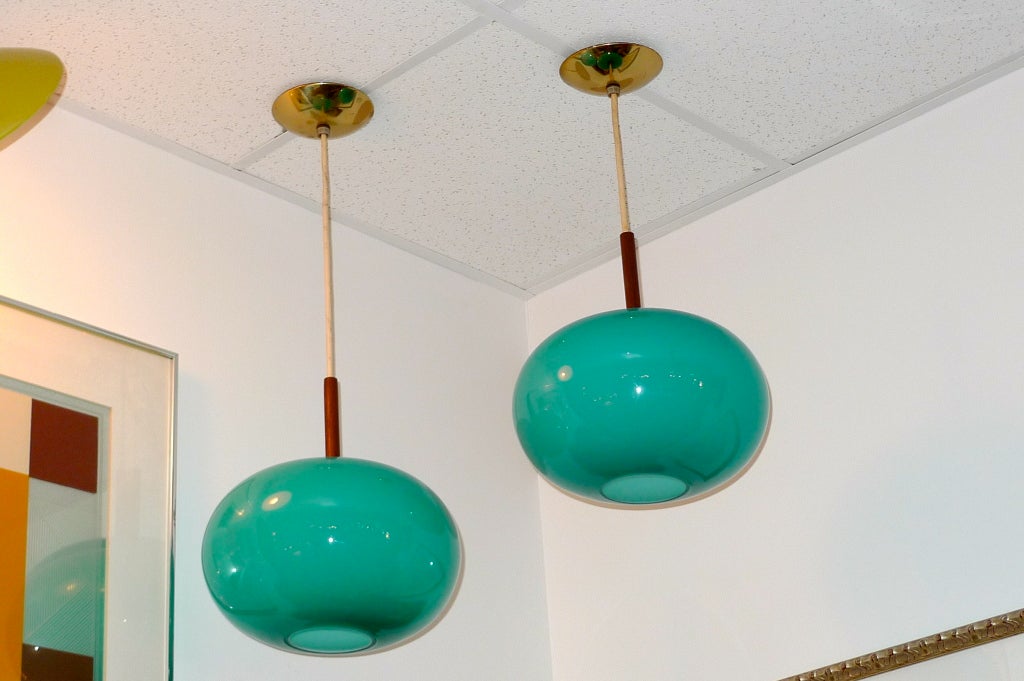 Pair of turquoise or seafoam colored glass pendants in shape of oblate spheroid by Prescolite of California.  Each hangs from vinyl coated wire and has a dark wood stem (possibly walnut or teak) and original brass ceiling caps (5.5