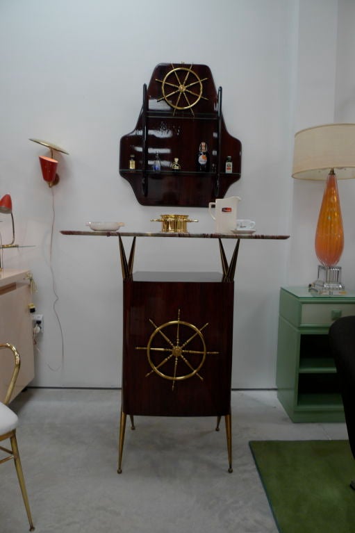 Gio Ponti employed this nautical motif in his designs around the time of the Andrea Doria commission. This standing bar consists of a highly polished mahogany cabinet held by angular brass scaffolds which support the curved marble top. The wall