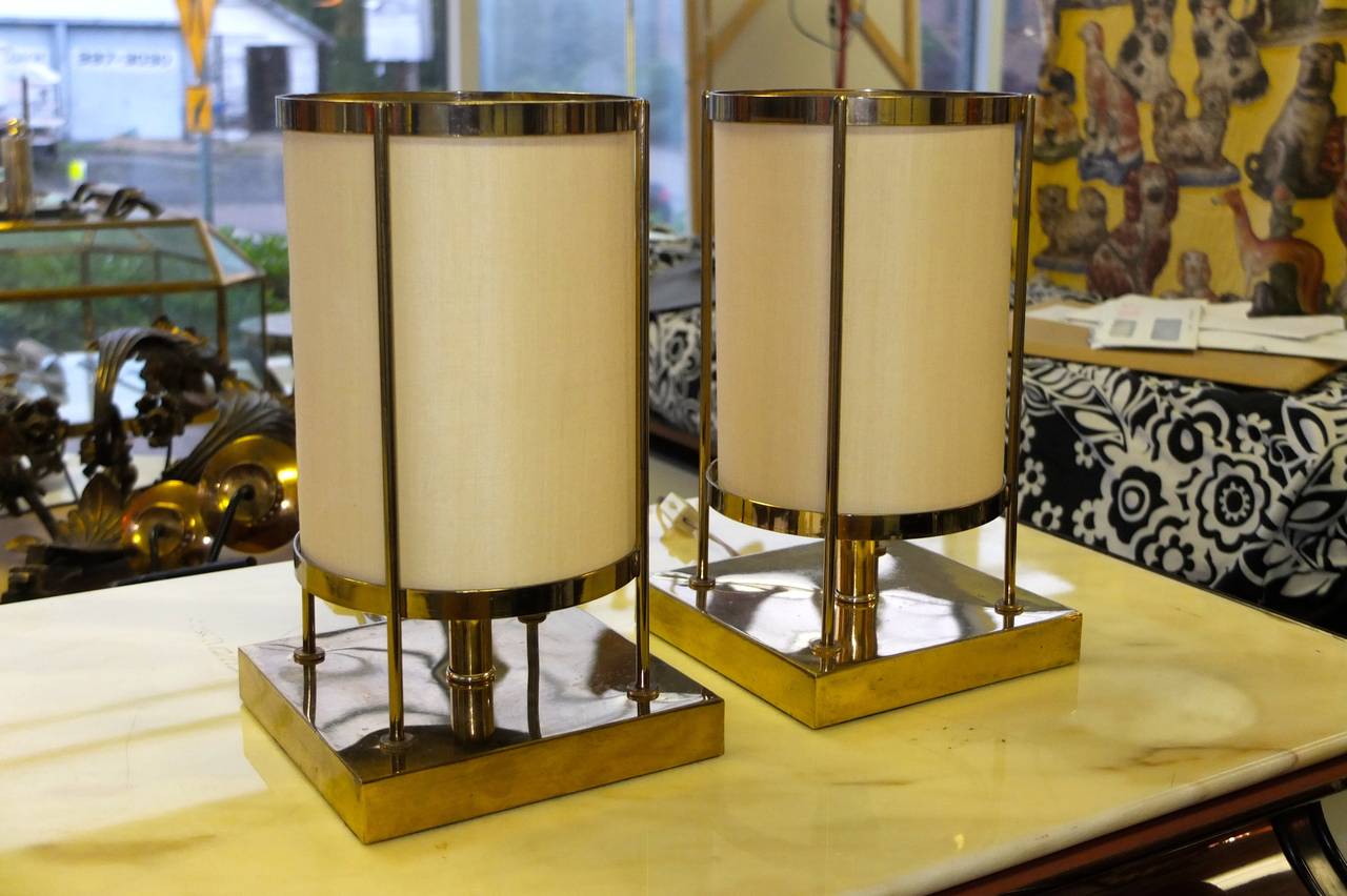 Pair of brass lantern lamps in the style of T. H. Robsjohn-Gibbings. 

Square brass bases with a four post drum structure supporting a custom-made cream colored cylinder shade around a single Edison porcelain screw socket bulb.

On off switch on